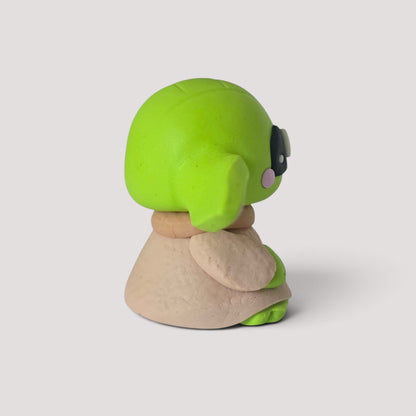 Statuetta artigianale El Muñequero Art di un personaggio verde in stile chibi con mantello beige, vista laterale
